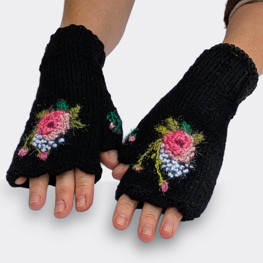 Mitaines pour femme noires présentées sur des mains à plat, avec une rose crochetée au centre de chaque composition florale. Les pétales roses en relief sont entourés de petits points blancs et de boutons floraux.