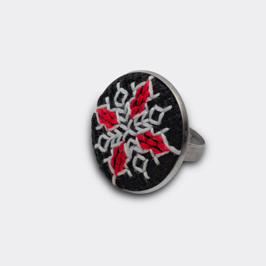 Motif brodé rouge et blanc appliqué sur tissu panama noir. Le plateau rond encadre la broderie graphique. Une bague brodée simple à porter.