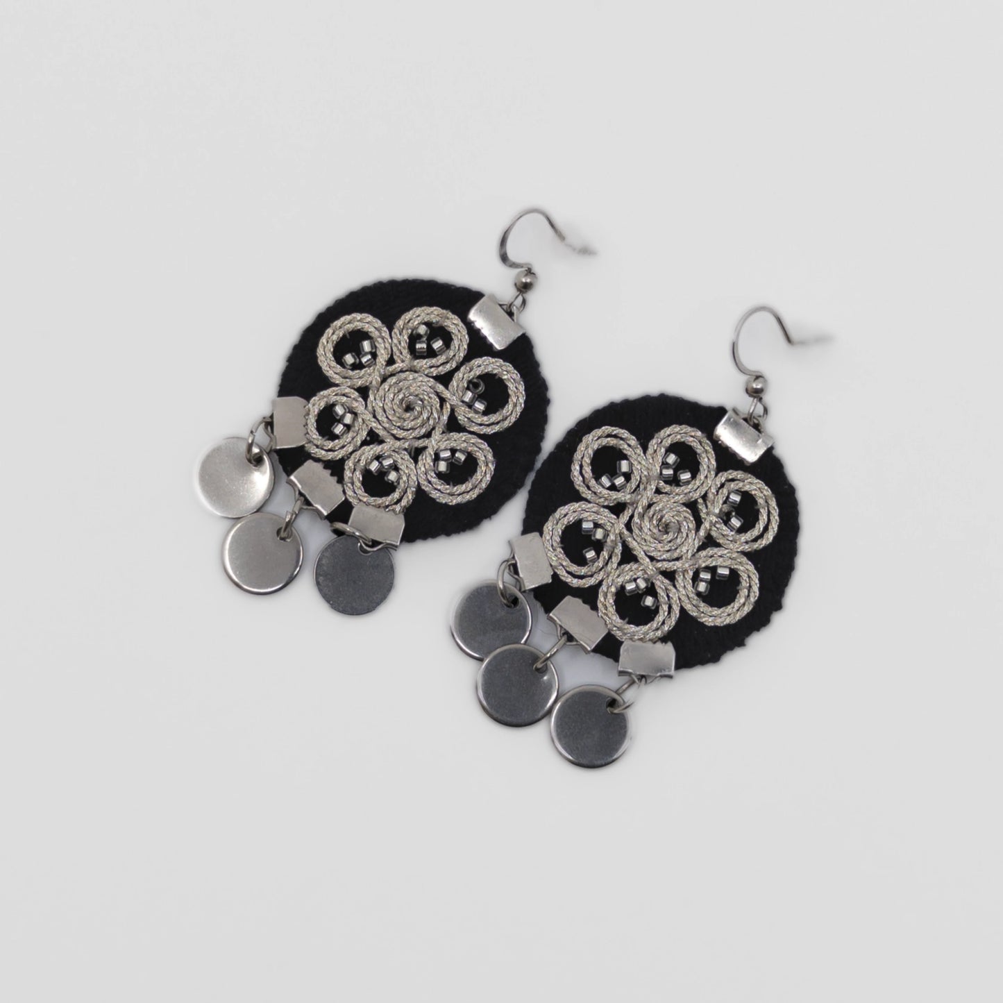 Boucles d’oreilles brodées au fil argenté sur tissu noir, motif spiralé inspiré du folklore serbe. Bijou textile réalisé à la main selon la technique zlatovez. Vue produit sur fond neutre.