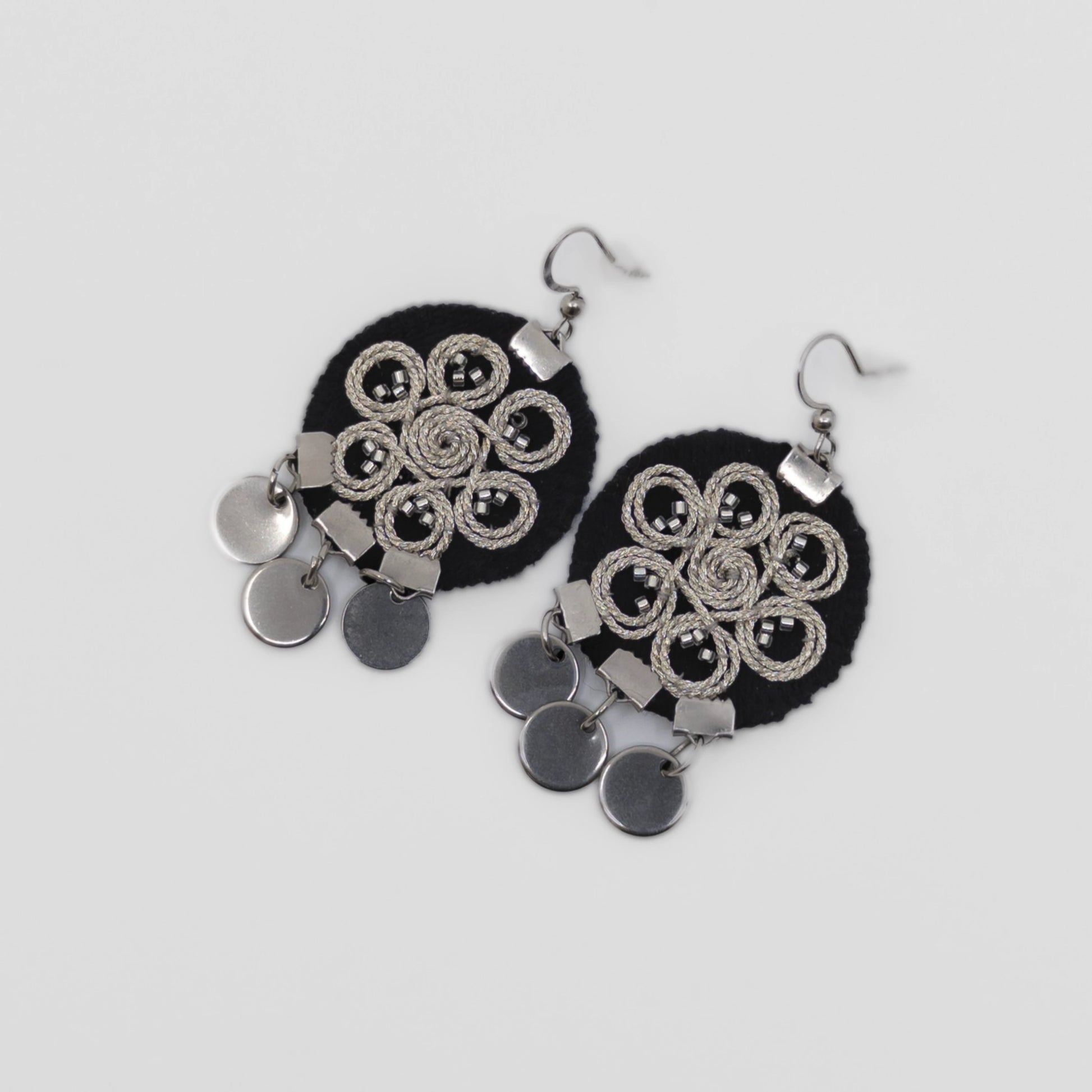 Boucles d’oreilles brodées au fil argenté sur tissu noir, motif spiralé inspiré du folklore serbe. Bijou textile réalisé à la main selon la technique zlatovez. Vue produit sur fond neutre.