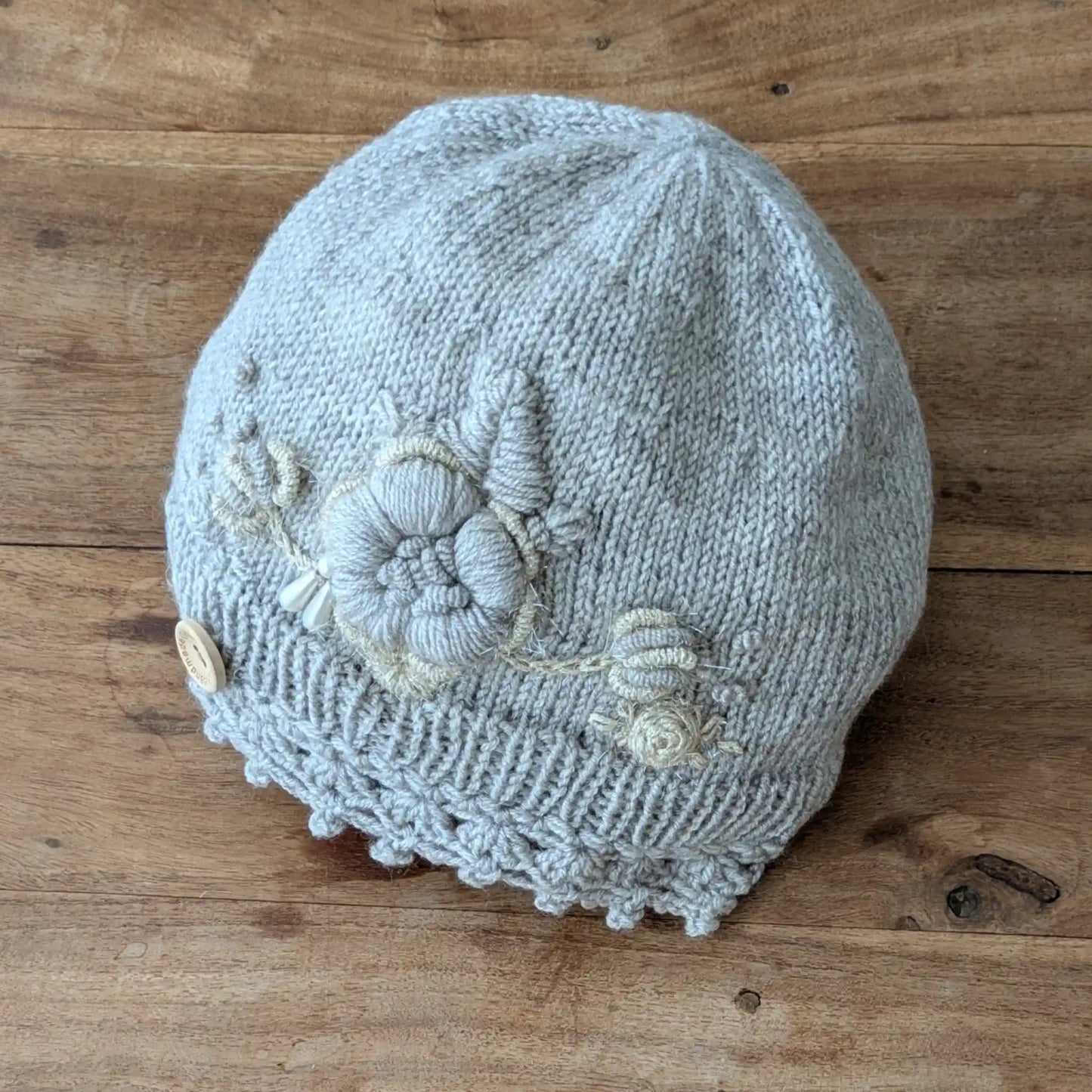 Bonnet gris clair tricoté main avec bord festonné, fleurs écrues et petits boutons décoratifs.