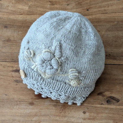 Bonnet gris clair tricoté main avec bord festonné, fleurs écrues et petits boutons décoratifs.