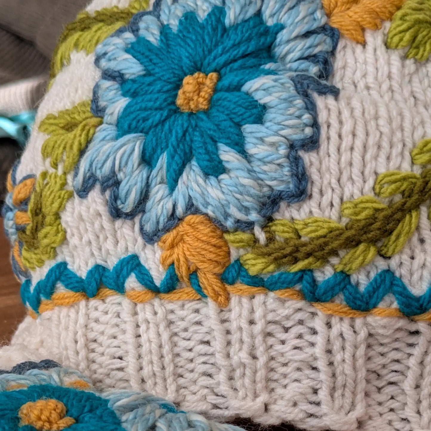 Gros plan sur le décor floral bleu et turquoise appliqué sur l’ensemble le bonnet assorti aux mitaines.