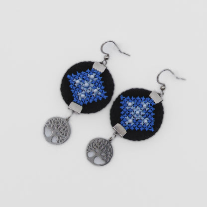 Boucles d’oreilles artisanales brodées en bleu sur fond noir, décor textile populaire et attaches en acier antiallergique.
