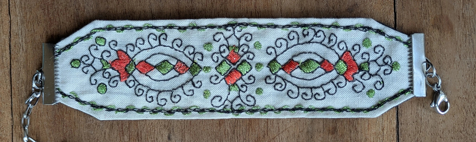 Détail d'un bracelet entièrement brodé à la main, écru, avec quelques pointes vertes et rouges-oranges ci et là. Fermoir ajustable en acier.