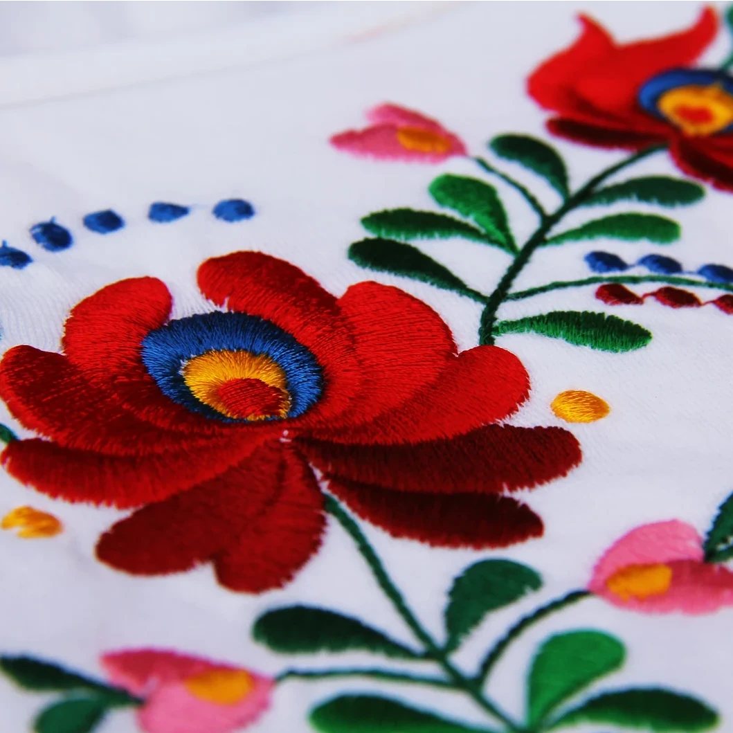 Vue en gros plan de la broderie de ce t shirt blanc à manches courtes. On distingue bien la fleur rouge typique des broderies Matyó entourée de délicates feuilles vertes.