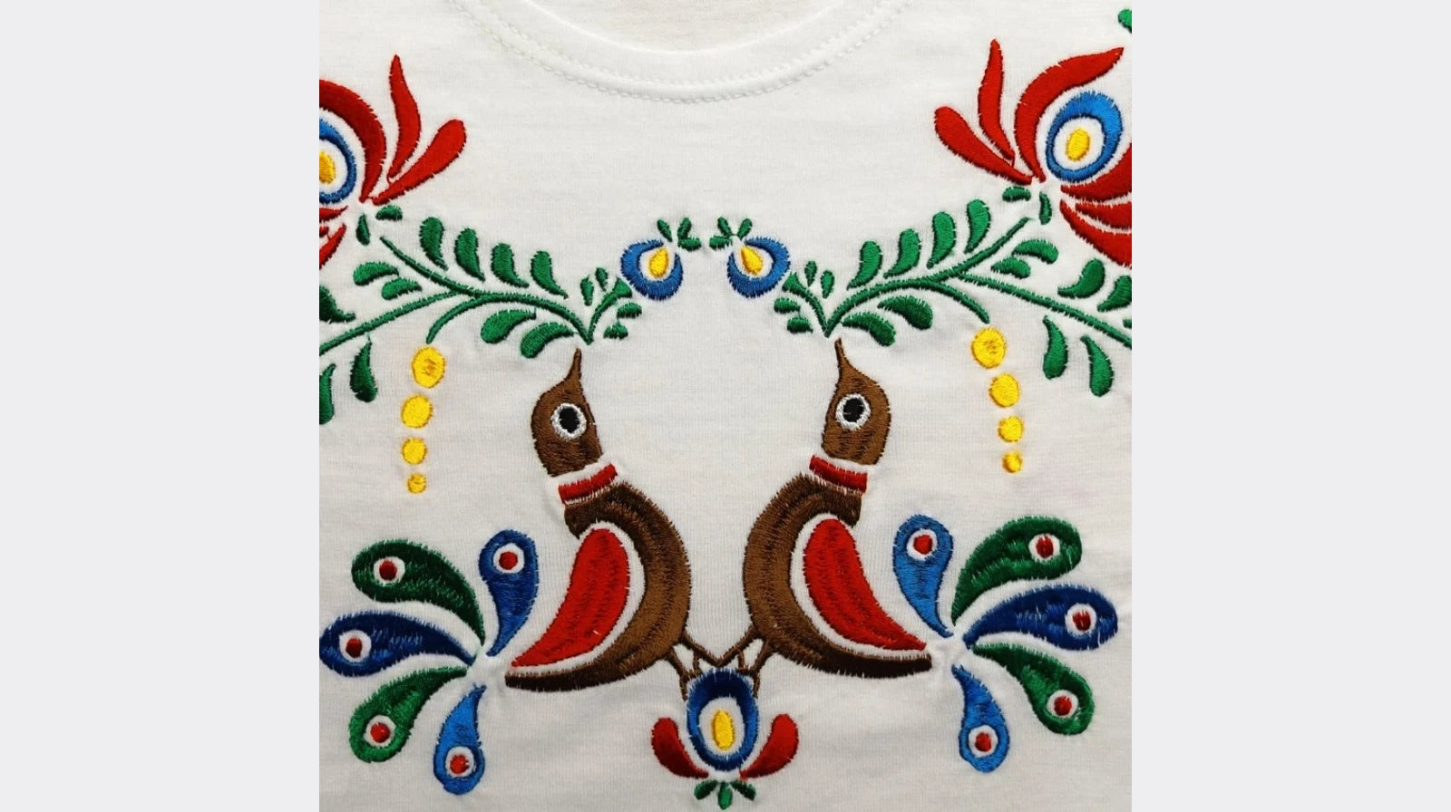 Détail de la broderie du tshirt pour enfant. Deux oiseaux multicolores entourés de fleurs et de feuilles se font face.