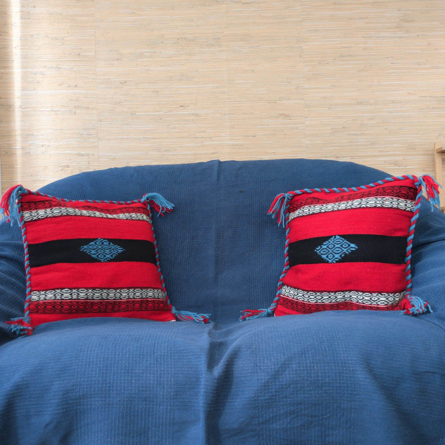 Visu de deux coussins sur un canapé avec une housse rouge, noire et bleue en laine tissée à la main.
Les housses ont une tresse et des pompons aux coins