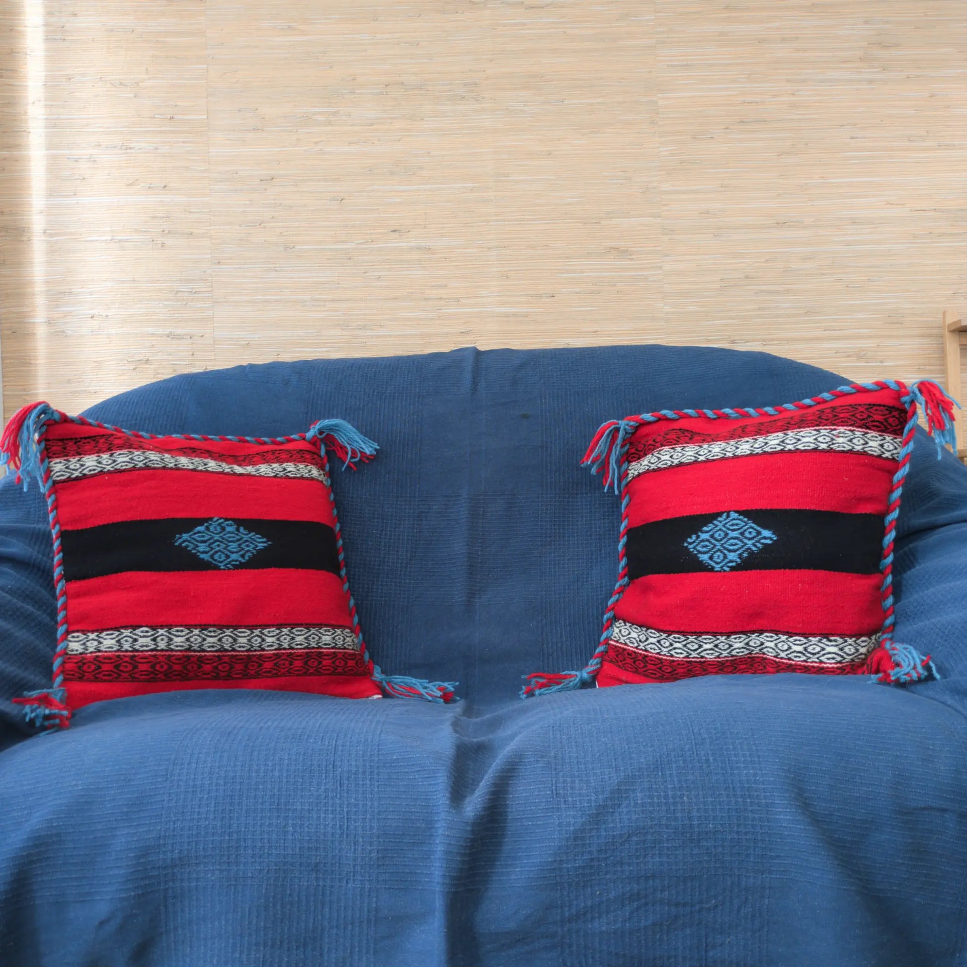 Visu de deux coussins sur un canapé avec une housse rouge, noire et bleue en laine tissée à la main.
Les housses ont une tresse et des pompons aux coins