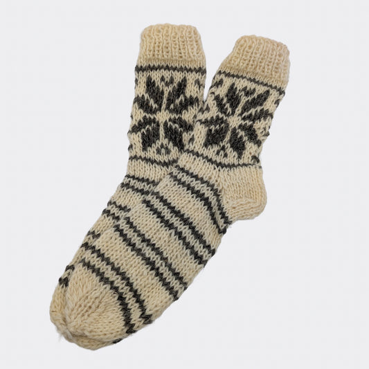 Paire de chaussettes en laine tricotées à la main, présentée à plat. Le fond blanc naturel est parcouru de rayures et de motifs géométriques gris. La maille épaisse et le bord côtelé sont visibles.