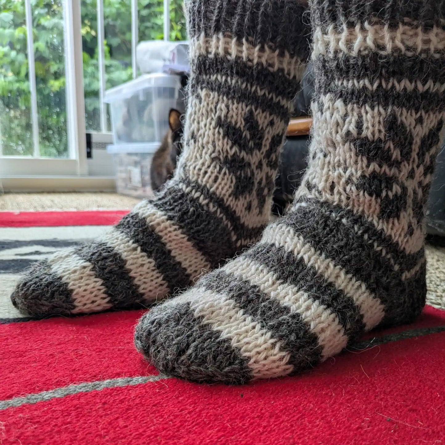 Paire de chaussettes en laine grise portée en intérieur. Les rayures du pied contrastent avec les motifs écrus de la tige. Tricot rustique et épais.