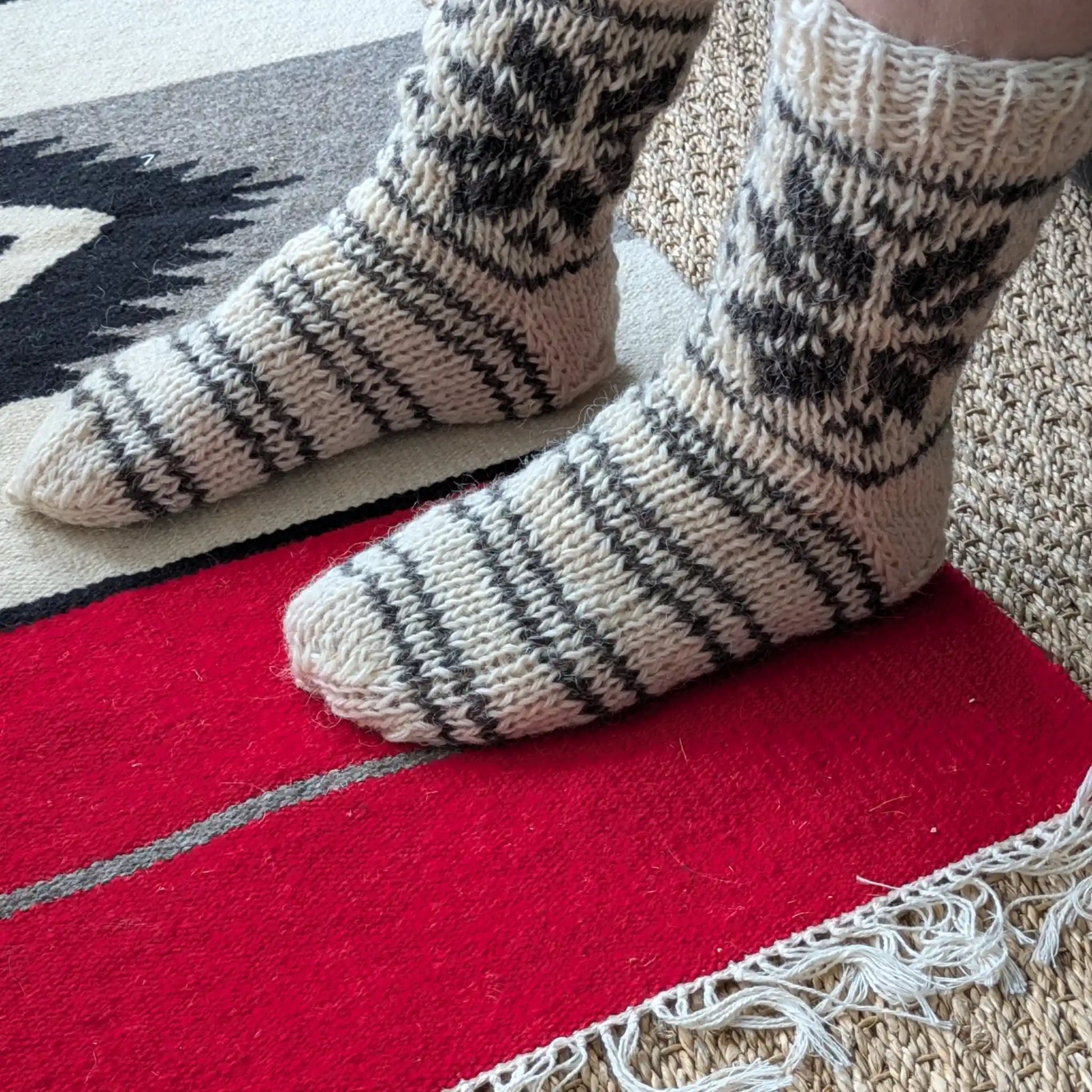 Chaussettes en laine épaisses portées en intérieur. Les rayures grises et blanches suivent le dessus du pied, tandis que les motifs géométriques couvrent la tige. Le talon uni apparaît sur le côté.