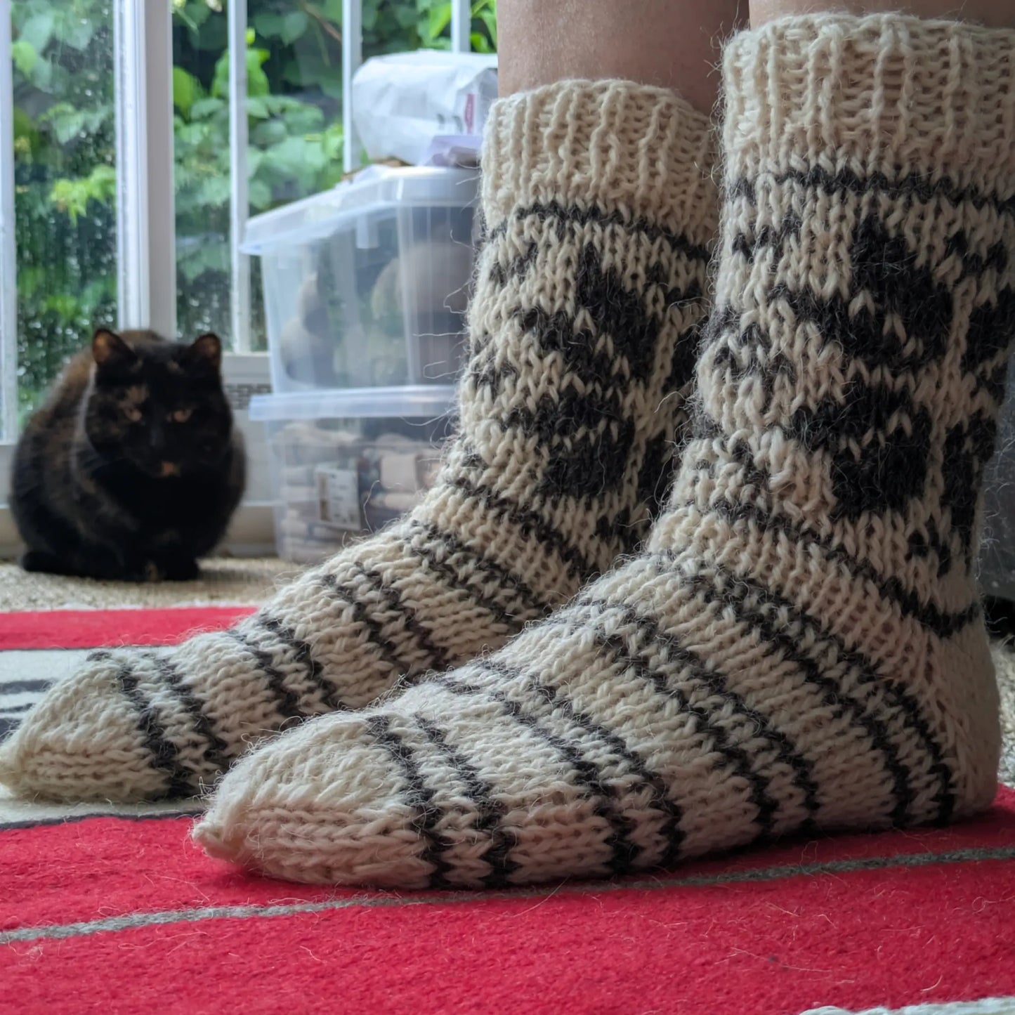 Chaussettes en laine tricotées à la main portées en gros plan. Les rayures grises couvrent le pied et les motifs géométriques remontent sur la tige. Un chat noir apparaît à l’arrière-plan.