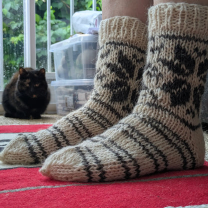 Chaussettes en laine tricotées à la main portées en gros plan. Les rayures grises couvrent le pied et les motifs géométriques remontent sur la tige. Un chat noir apparaît à l’arrière-plan.