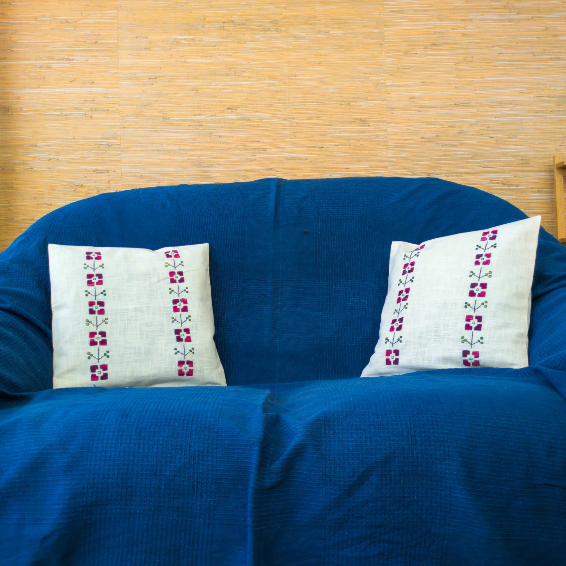 Canapé décoré d’un coussin en lin brodé de fleurs rouges géométriques. Les colonnes florales se distinguent nettement. Une ambiance artisanale douce.