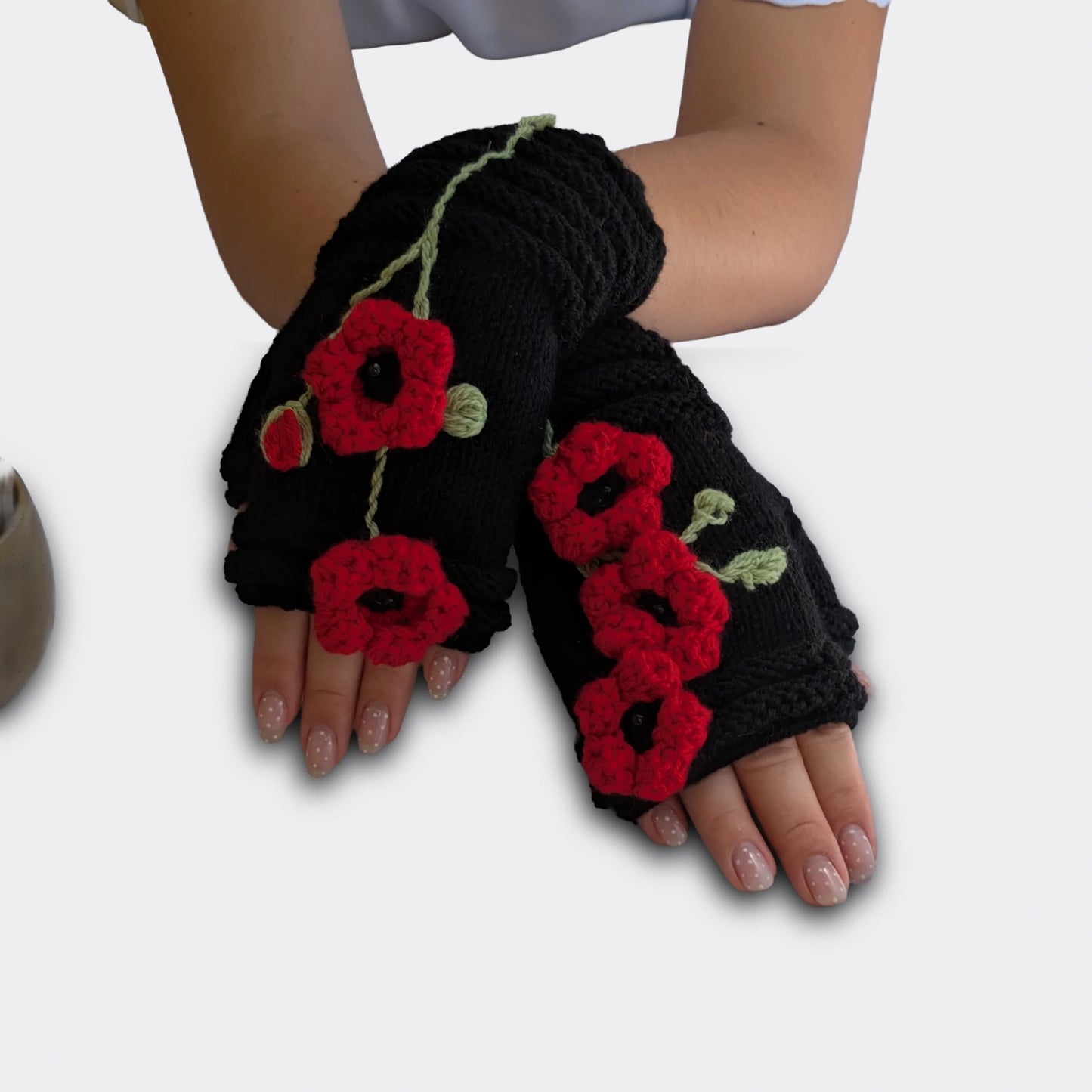Mitaines noires portées mains croisées, tricotées main avec coquelicots rouges appliqués et tiges vert clair brodées.