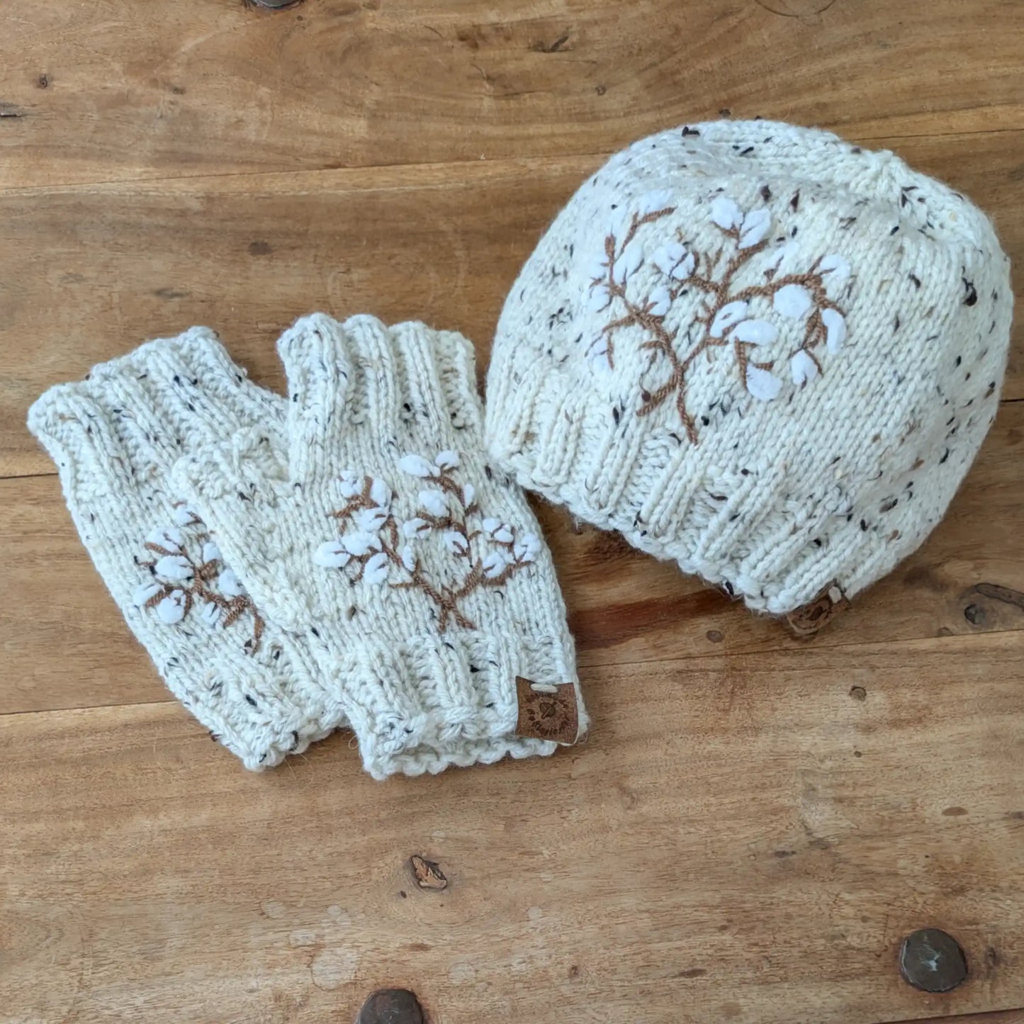 Bonnet et mitaines assortis écru moucheté posés à plat, décorés de branches de coton stylisées.