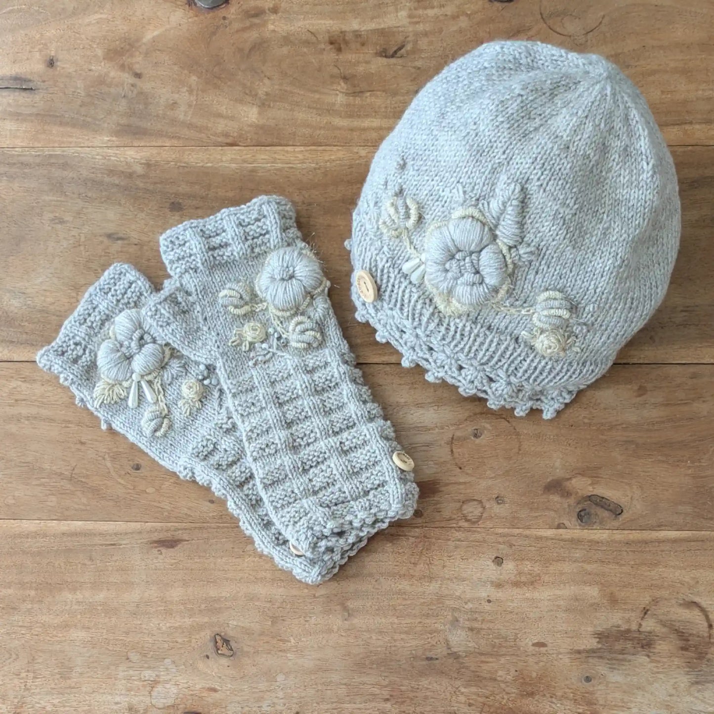 Bonnet et mitaines assortis gris clair posés à plat, décorés de fleurs écrues et de détails en relief.