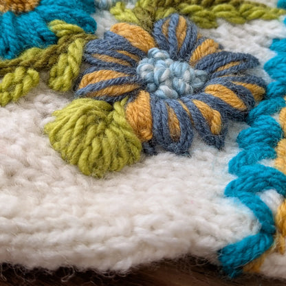 Gros plan sur les fleurs bleues et jaunes en relief de ces gants sans doigts blanc cassé.