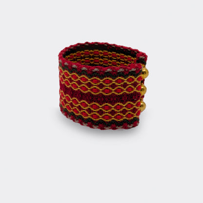 Bracelet tissé rouge et or à motifs géométriques inspirés du folklore des Balkans. Les fils de coton et le fil doré créent une trame lumineuse, vue de face.