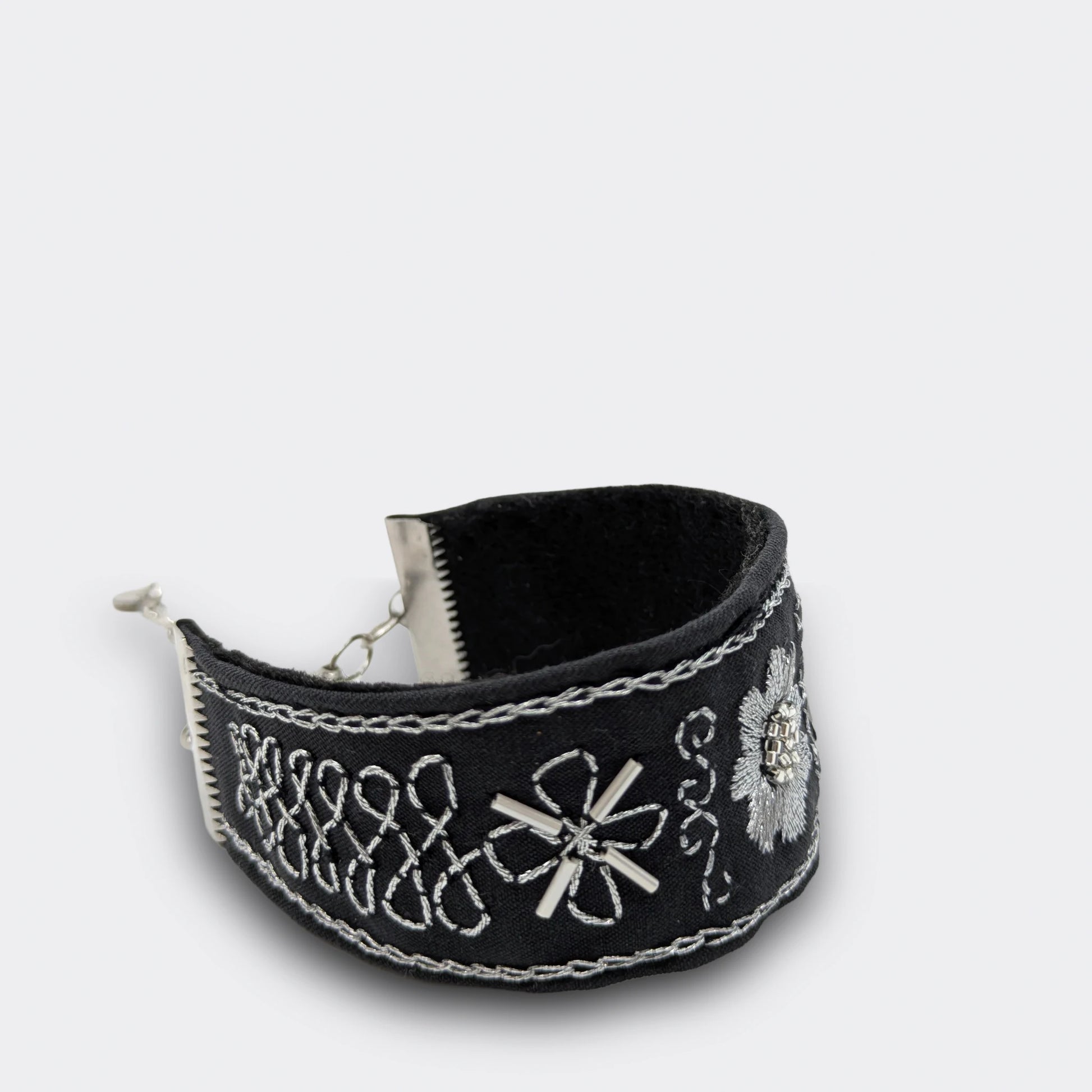 Gros plan sur la broderie argentée d’un bracelet textile noir, ornée de lignes et volutes en points traditionnels. Fait main en Serbie, détail du motif.