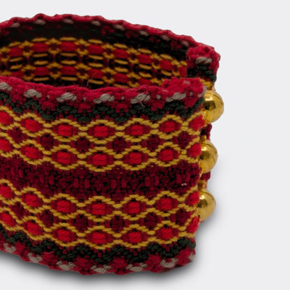 Gros plan sur la texture du bracelet tissé rouge et or. On distingue les entrelacs de fils et la finesse du tissage manuel