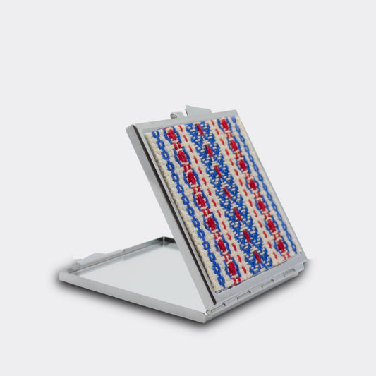 Miroir de poche décoré d’un tissage rouge et noir sur fond écru. Le boîtier est ouvert et laisse voir la surface du miroir. Accessoire léger et discret.