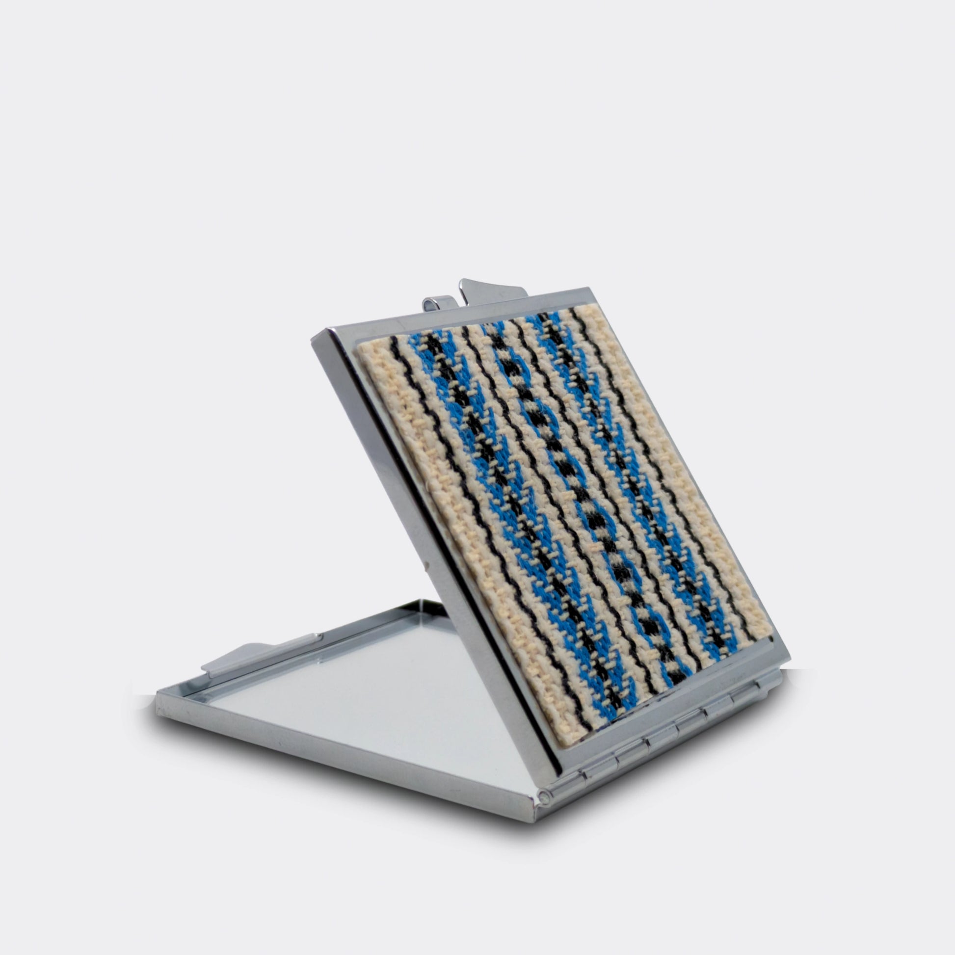 Miroir de poche décoré d’un tissu tissé bleu et noir sur fond écru. Le boîtier métallique est ouvert, révélant le miroir. Un accessoire fin et facile à glisser dans un sac.