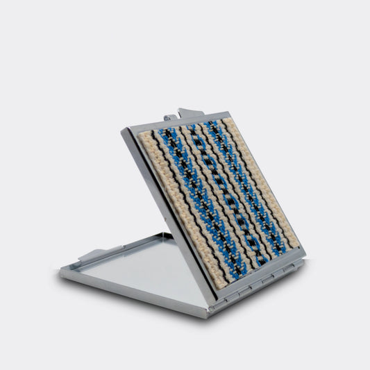 Miroir de poche décoré d’un tissu tissé bleu et noir sur fond écru. Le boîtier métallique est ouvert, révélant le miroir. Un accessoire fin et facile à glisser dans un sac.