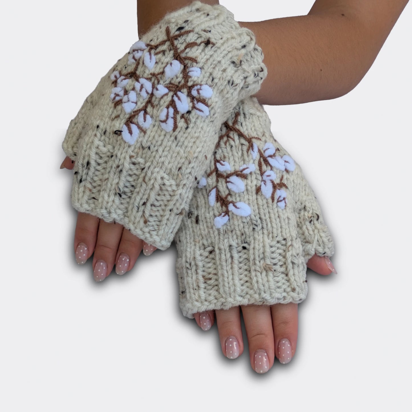 Mitaines brodées écrues portées mains croisées. Le motif montre des branches de coton stylisées, avec fleurs blanches et tiges brunes sur fond moucheté. La maille présente un discret effet tweed.