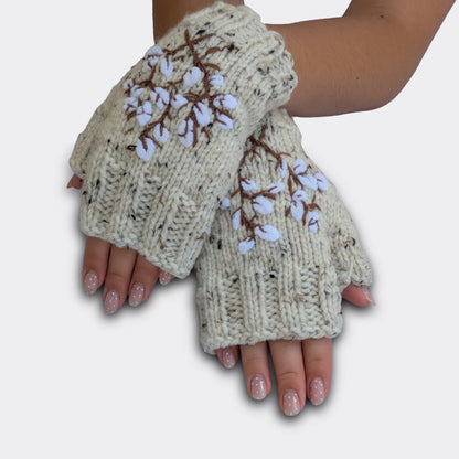 Mitaines brodées écrues portées mains croisées. Le motif montre des branches de coton stylisées, avec fleurs blanches et tiges brunes sur fond moucheté. La maille présente un discret effet tweed.