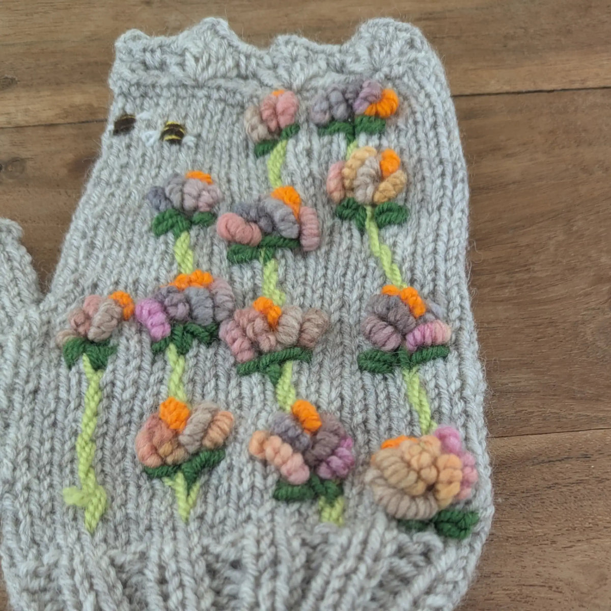Détail des fleurs en relief sur des mitaines grises artisanales. Les petites corolles aux nuances de rose, mauve, pêche et orange vif se succèdent sur plusieurs tiges vert tendre.