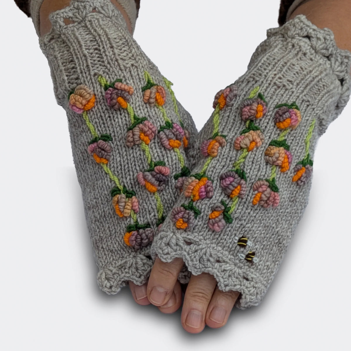 Des mains légèrement croisées portent des mitaines artisanales grises à fleurs en relief. Les petites fleurs multicolores se succèdent le long de tiges vert tendre, accompagnées de motifs d'abeilles.