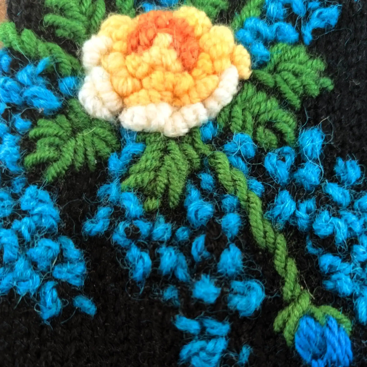 Vue en gros plan de la fleur centrale crochetée sur une paire de mitaines artisanale. On y voit également quelques points bleus et du feuillage.