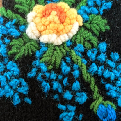 Vue en gros plan de la fleur centrale crochetée sur une paire de mitaines artisanale. On y voit également quelques points bleus et du feuillage.