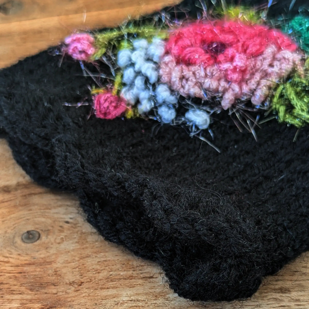 Vue en détail du bord supérieur festonnée d'une paire de mitaine noire pour femme. Elle est ornée d'une rose aux teintes rosées, de tiges vert olive et de petits points blancs.