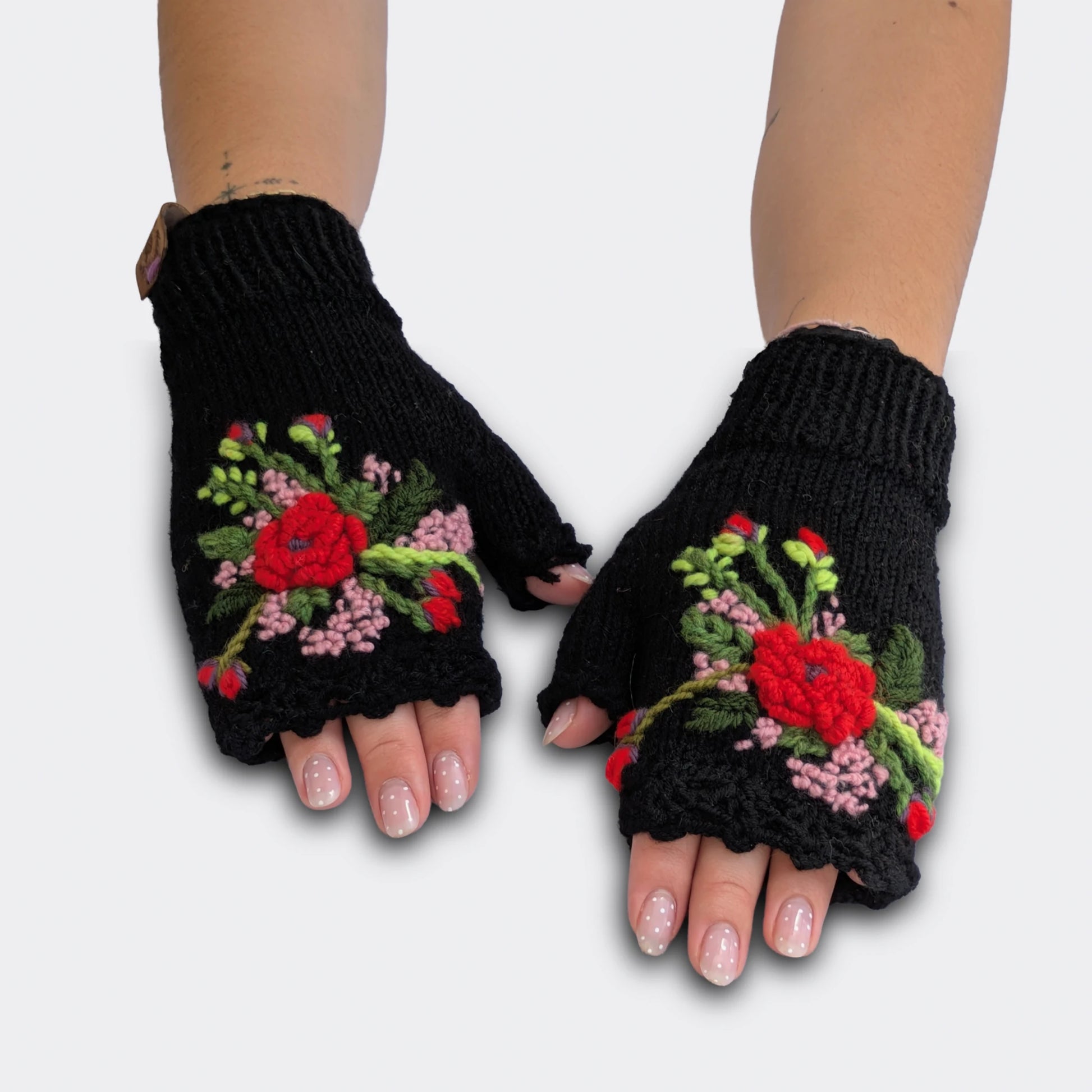 Mitaines noires tricotées main, portées à plat. Le décor floral associe des fleurs rouges et rose pâle appliquées en relief sur la maille jersey. Le poignet présente un bord en côtes et le haut une finition festonnée.
