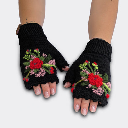 Mitaines noires tricotées main, portées à plat. Le décor floral associe des fleurs rouges et rose pâle appliquées en relief sur la maille jersey. Le poignet présente un bord en côtes et le haut une finition festonnée.
