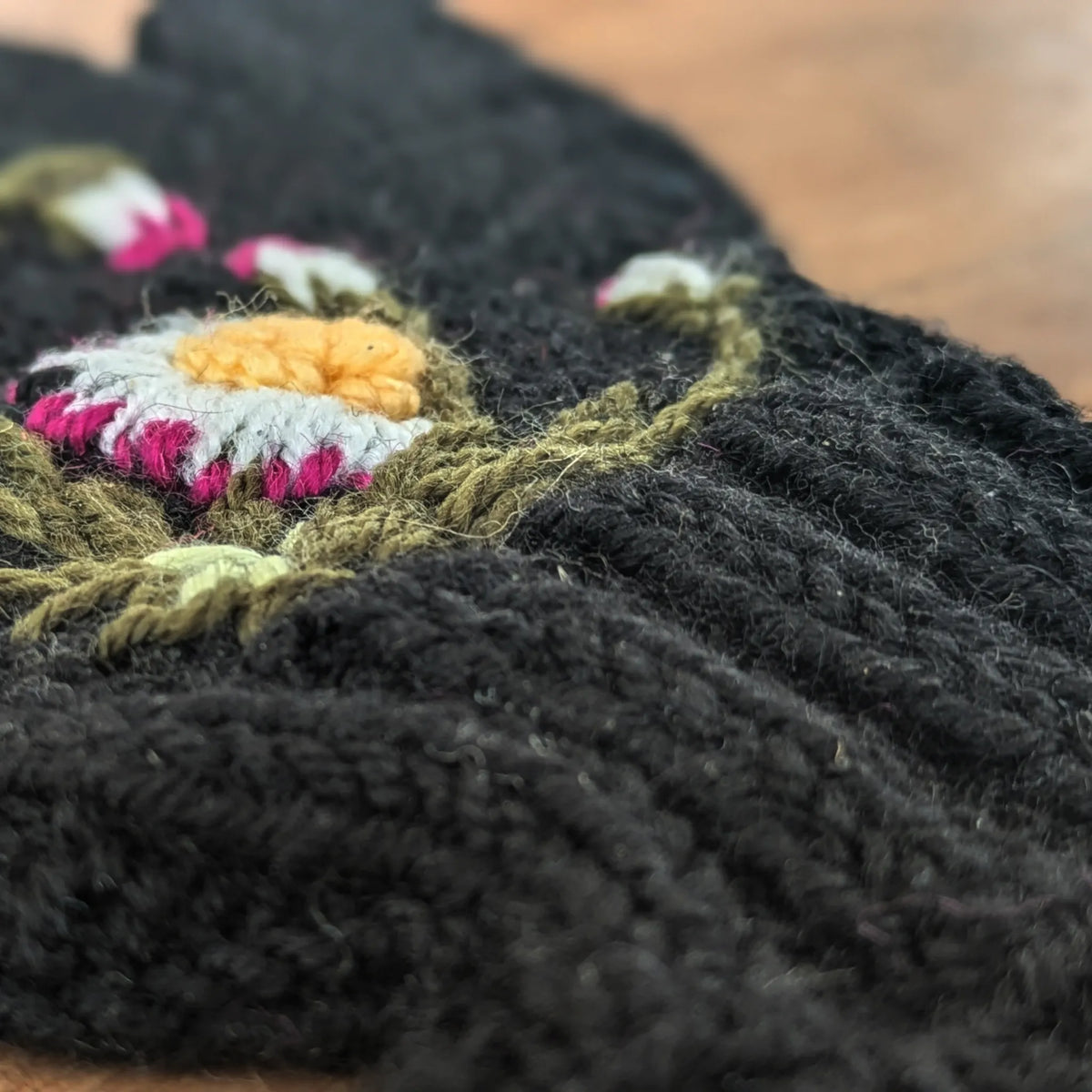 Détail d'une fleur crochetée jaune et blanche avec sa tige vert olive sur une mitaine noire dont on voit aussi le bord côte.