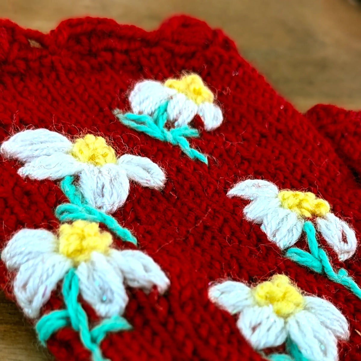 Vue en biais d'une paire de mitaines posée à plat. On y voit 5 fleurettes brodées sur le dessus contrastant avec le rouge profond des mitaines faites main en Serbie.