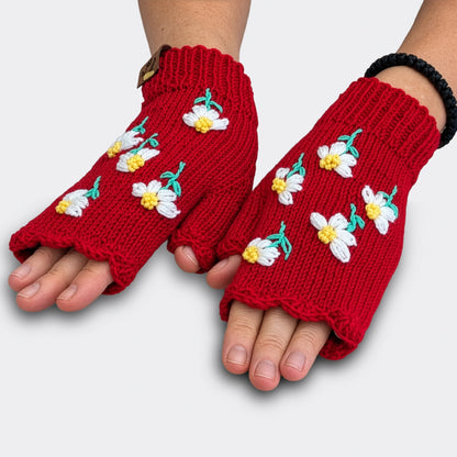 Sur ces mitaines rouges tricotées à la main, des fleurs blanches aux tiges vertes apparaissent séparément. La vue à plat laisse voir les petits motifs floraux et le bord supérieur festonné.