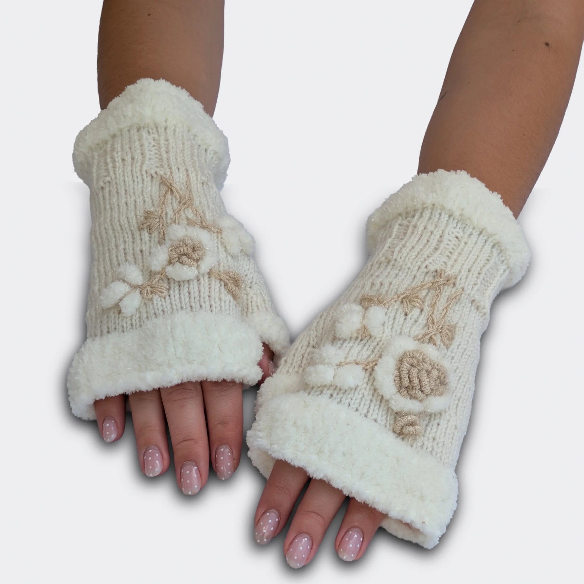 Vue portée à plat de mitaines artisanales blanches. Le décor floral appliqué associe fleur en relief, petites boules blanches et tige champagne légèrement lumineuse. Les bords pelucheux encadrent le poignet et le haut de la main.