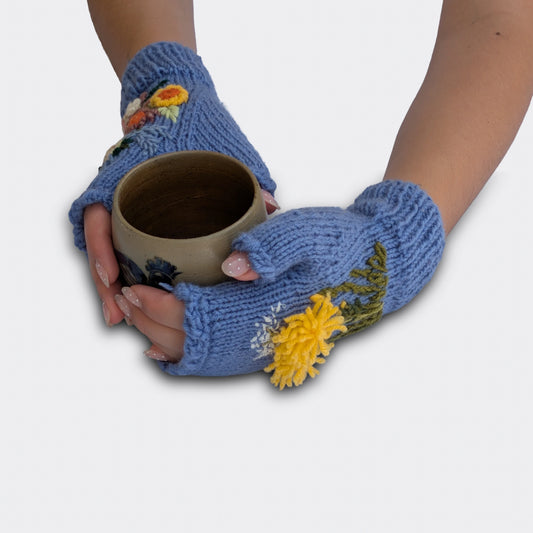 Mitaines bleu lavande tenant une tasse, avec pissenlit jaune brodé, fleurs crème et orange, et bord côtelé au poignet.