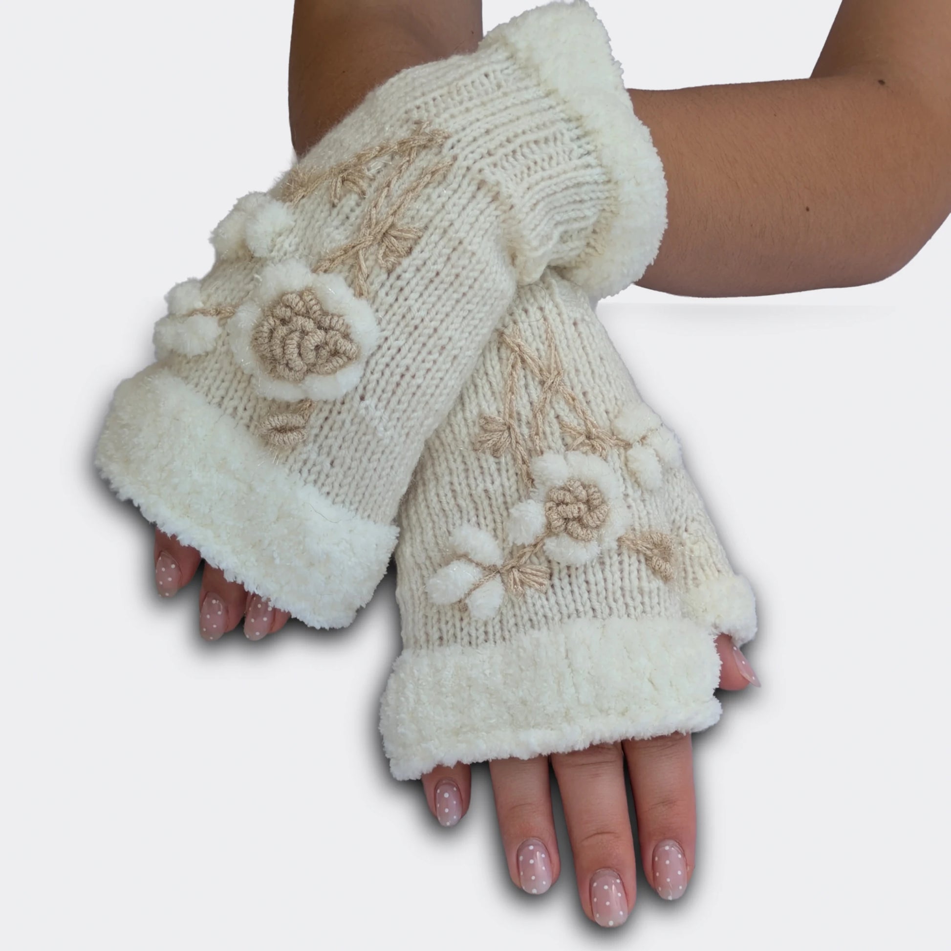 Mitaines brodées blanches portées mains croisées. La fleur stylisée en relief ressort sur une maille tricotée main, avec de petites boules blanches façon flocons. Le haut de la mitaine et le pouce présentent un ourlet pelucheux.