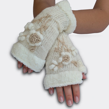 Mitaines brodées blanches portées mains croisées. La fleur stylisée en relief ressort sur une maille tricotée main, avec de petites boules blanches façon flocons. Le haut de la mitaine et le pouce présentent un ourlet pelucheux.