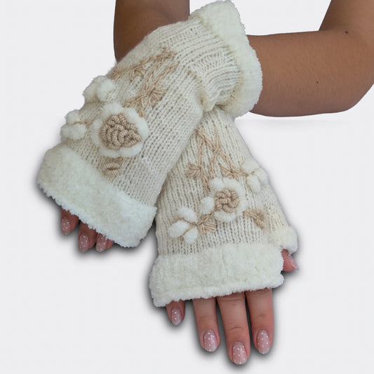 Mitaines brodées blanches portées mains croisées. La fleur stylisée en relief ressort sur une maille tricotée main, avec de petites boules blanches façon flocons. Le haut de la mitaine et le pouce présentent un ourlet pelucheux.