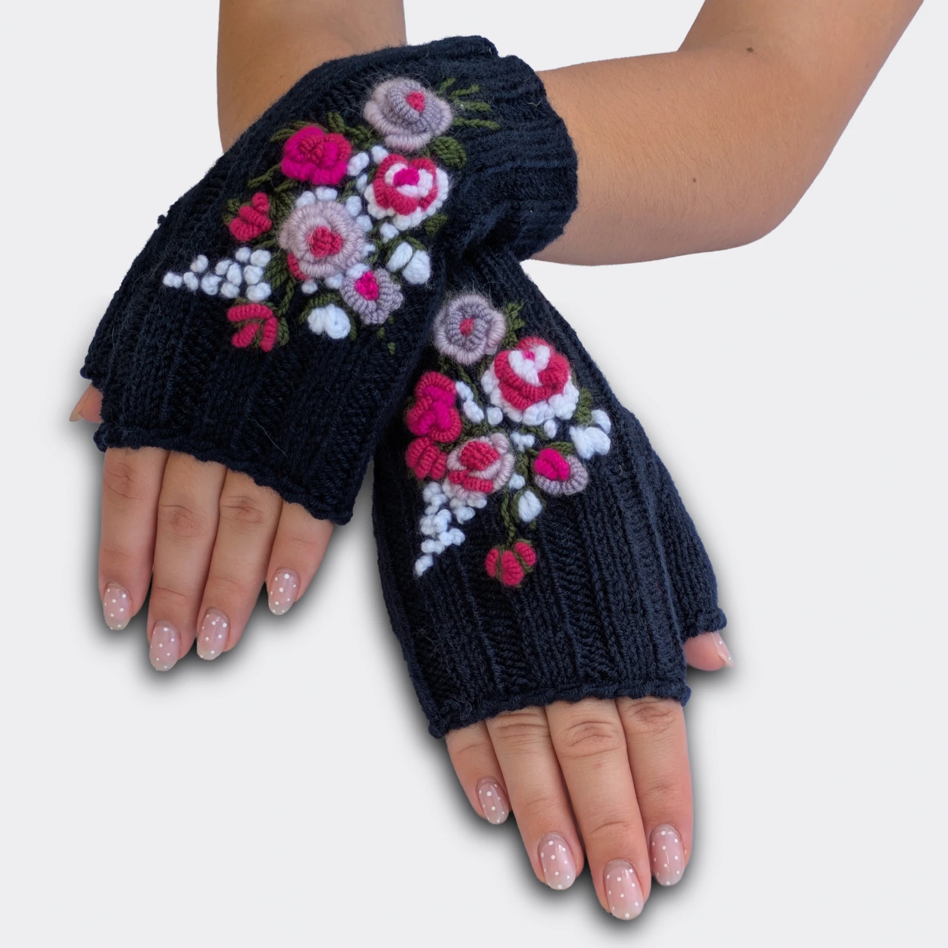 Mitaines brodées bleu nuit portées mains croisées. Le motif floral associe roses crochetées, petites fleurs blanches et feuillage vert brodé. La maille en côtes donne une texture souple sur toute la paire.