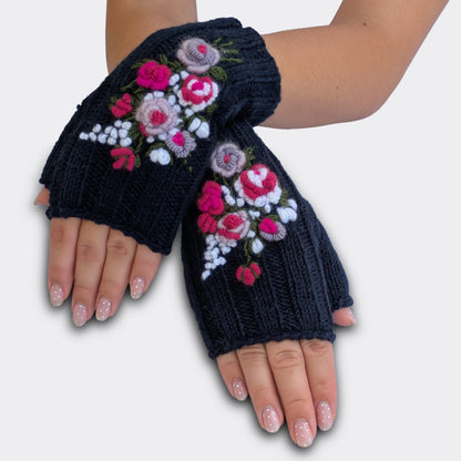 Mitaines brodées bleu nuit portées mains croisées. Le motif floral associe roses crochetées, petites fleurs blanches et feuillage vert brodé. La maille en côtes donne une texture souple sur toute la paire.