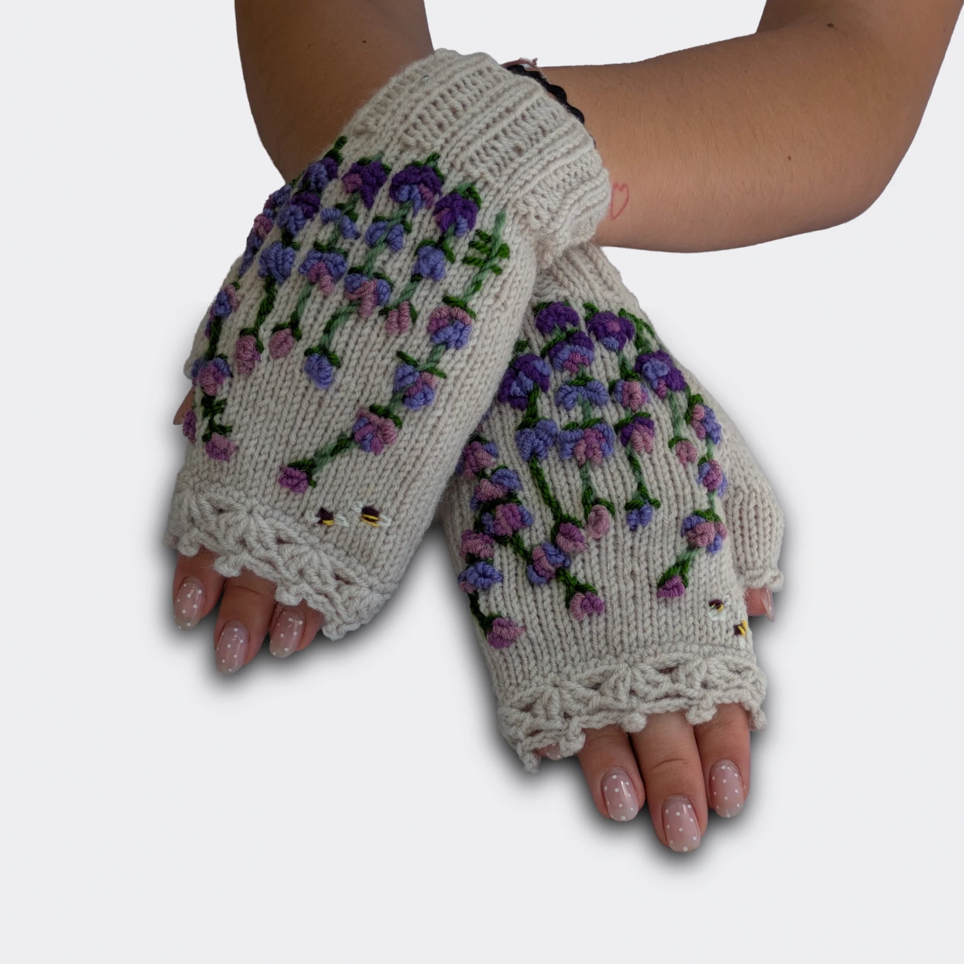 Mitaines brodées gris clair portées mains croisées. Le motif floral butiné associe petites fleurs mauves, feuillage vert et deux abeilles sur une base en maille claire. Le poignet présente un bord-côte.
