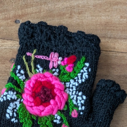 Gros plan sur la fleur rose en relief, le feuillage vert et les broderies blanches sur des mitaines noires.